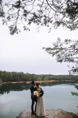 The bride and groom stand on a stone near the lake in the forest and kiss. The bride holds a wedding bouquet in boho style. Wedding in the forest.