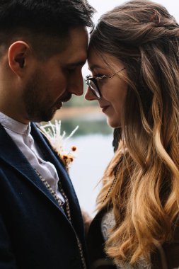 The bride and groom in close-up profile touch their foreheads and smile. The bride has long blond hair styled in curls with feathers.