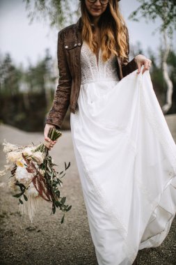 Bride with light curls in a lace dress, brown leather jacket and glasses. The girl walks through the forest, holds the bride's bouquet in the boho style and the hem of the dress.