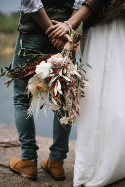 The bride and groom standing by the lake with their backs. The groom holds the bride's bouquet in boho style with peonies, feathers, and ears of corn. The bride in a long dress holds his hand.