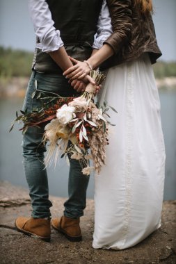 The bride and groom standing by the lake with their backs. The groom holds the bride's bouquet in boho style with peonies, feathers, and ears of corn. The bride in a long dress holds his hand.