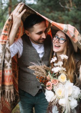 Bride and groom in boho style, laughing in the forest. The groom covers their head with a Navajo blanket. The bride holds a wedding bouquet of peonies, roses, feathers, and cereals.