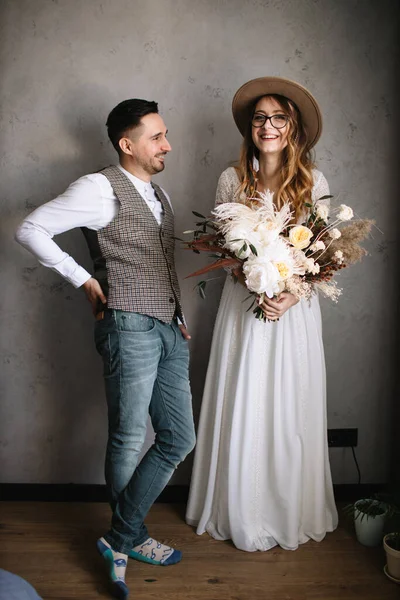 The bride and groom are standing and laughing. The bride in a white dress, hat, glasses, with feathers in her hair holds a bouquet in boho style and laughs. The groom looks at the girl and smiles.