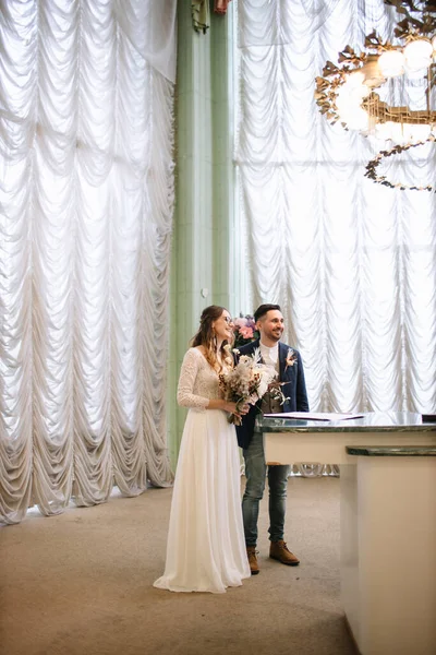 The bride and groom are standing at the registration desk in the registry office and laughing. The bride is holding a wedding bouquet. In the background are high windows with curtains and a chandelier