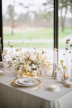 Düğün ziyafeti masasının süsü. Masanın ortasında bir buket beyaz ve bej karanfil, altın ve beyaz tabak, altın çatal bıçak, şarap kadehi, yanan mumlar var.. 