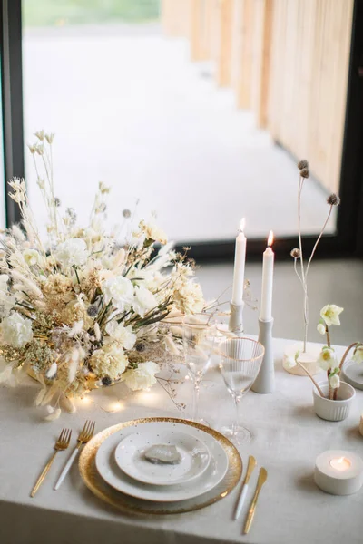 Serving and decoration of the wedding banquet table. On gold and white plates there is a place indicator, gold cutlery, wine glasses. In the background are flowers and burning candles.