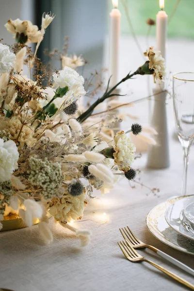 Banquet table decor in white and gold. Bouquet of carnations, wild flowers and grasses, golden cutlery, plates, wine glasses.