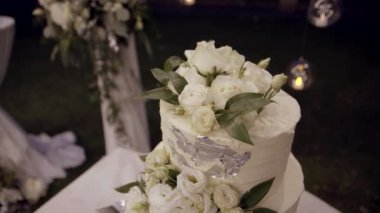 Düğün pastası, tören alanında yakın plan. Yaratıcı dekorlu beyaz ve pembe düğün pastası. Bahçe gece yapılacak bir düğün için dekore edilmiştir. Yavaş çekim.