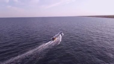 Bir sürat teknesinin şişme bir muz teknesini yüksek hızda çekerken görüntüsü. Video, tekneyi takip eden bir İHA tarafından çekildi. Deniz kenarında gezinti, deniz eğlencesi. 