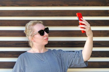 Sarışın bir kadın, parkta bir duvarın arkasında dikilirken ya da telefonla görüntülü konuşarak selfie çeker ya da iletişim kurar.