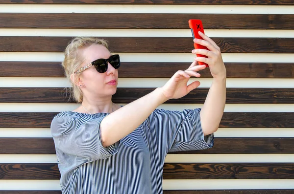 Sarışın bir kadın, parkta bir duvarın arkasında dikilirken ya da telefonla görüntülü konuşarak selfie çeker ya da iletişim kurar.