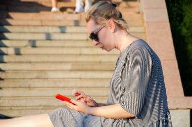 Bir kadın, bir yaz günü parkta otururken akıllı telefonundan internet alımları yapar ya da anında haberleşir.