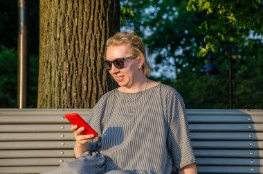 Bir kadın, bir yaz günü parkta otururken akıllı telefonundan internet alımları yapar ya da anında haberleşir.