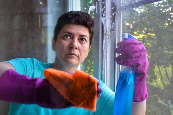 Kısa siyah saçlı, lastik eldivenli bir kadın elinde cam temizleyicisi tutarken evinin camını temizliyor. Temizlik hizmetleri, cam yıkama, ev işi.