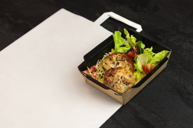 Siyah arka planda izole edilmiş beyaz torba üzerinde eko kağıt kabın içinde tavuk pirzolası kuskus, marul ve domates bulunan sağlıklı bir kase. Sağlıklı yemek servisi ve günlük istihkak konsepti. Yakın plan..