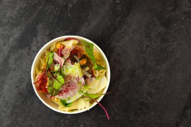 Kabuklu makarna, krema sosu, parmesan peyniri ve yeşillikler koyu arka planda yuvarlak kağıt yemek kabında. Paket servis ve sağlıklı yemek teslimatı kavramı. Metin için boş alan.