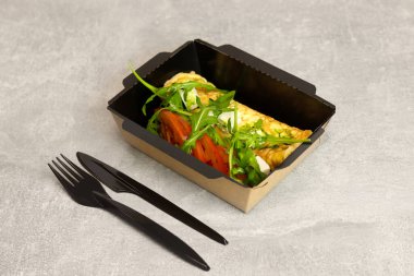 Fransız omleti, domates ve roka salatası siyah çatal ve bıçakla kaplanmış gri bir kapta. Kahvaltıda. Sağlıklı yemek teslimatı kavramı.