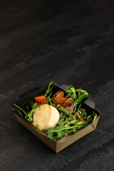 Savory souffle salmon and salad with arugula, tomatoes and green beans in black paper takeaway box on dark background. Healthy food delivery concept.