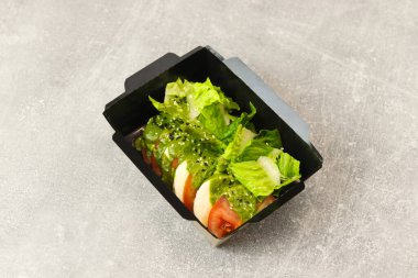 Mozarella soslu taze İtalyan caprese salatası ve gri arka planda siyah paket servis kutusunda domates. Sağlıklı yemek teslimatı kavramı.