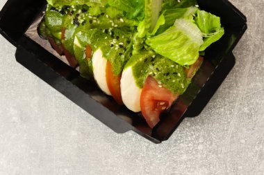 Mozarella soslu taze İtalyan caprese salatası ve gri arka planda siyah paket servis kutusunda domates. Sağlıklı yemek teslimatı kavramı.