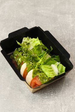 Mozarella soslu taze İtalyan caprese salatası ve gri arka planda siyah paket servis kutusunda domates. Sağlıklı yemek teslimatı kavramı.