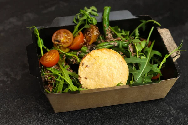 Savory souffle salmon and salad with arugula, tomatoes and green beans in black paper takeaway box on dark background. Healthy food delivery concept.