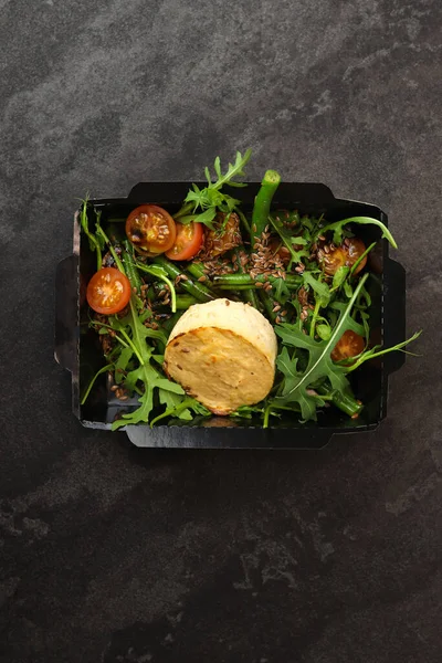 Savory souffle salmon and salad with arugula, tomatoes and green beans in black paper takeaway box on dark background. Healthy food delivery concept.