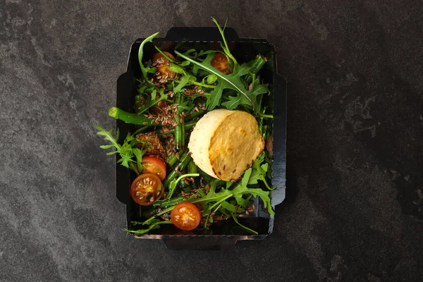 Savory souffle salmon and salad with arugula, tomatoes and green beans in black paper takeaway box on dark background. Healthy food delivery concept.