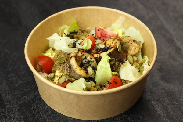 Fish salad. Fresh vegetable salad with baked fish fillet. Fish salad with fried fillet and fresh vegetables in eco paper bowl on dark background. Food delivery service and daily ration concept.