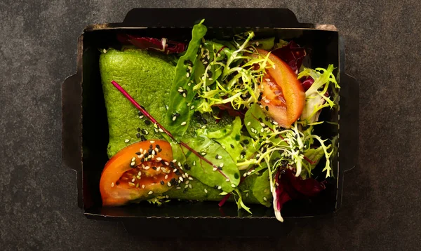 Spinach pancake with green lettuce, tomato and sesame salad in a black paper container on a dark background. Healthy food delivery service and daily ration concept.