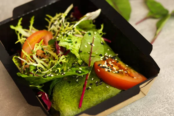 Spinach pancake with green lettuce, tomato and sesame salad in a black paper container on a gray background. Healthy food delivery service and daily ration concept.