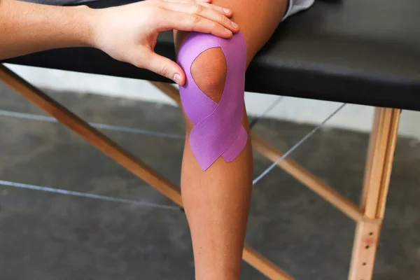 Fizyoterapist hastanın dizine kineziyoloji bandı uyguluyor. Genç bayan sporcuyu tedavi eden terapist. Kineziyoloji bandı. Travma sonrası rehabilitasyon, spor fizyoterapisi.