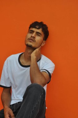 İyi görünümlü bir Hintli genç dışarıda oturmuş fotokopi aletiyle turuncu duvarlara poz veriyor. 