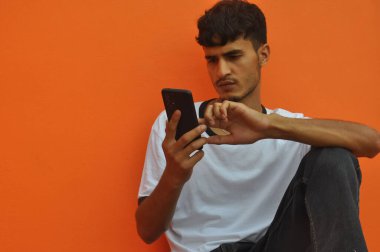 A handsome Indian guy using his phone while sitting against orange wall background with copy space 