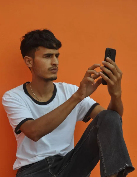 コピースペースとオレンジの壁の背景に座っている間彼の携帯電話を使用してハンサムなインド人 — ストック写真