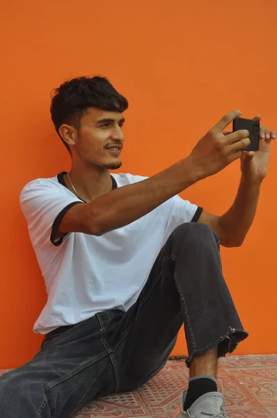 照片上的一个快乐的年轻人 一边拿着手机 一边靠着橙色墙的背景图片 一边带着复制的空间 — 图库照片