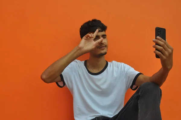コピースペースとオレンジの壁の背景に座って 自己撮影中に平和サインを示す若い男 — ストック写真