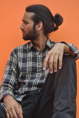 A good looking young man with beard and hair bun looking sideways while sitting outside, posing against orange wall background