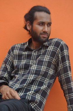 A good looking young man with beard and hair bun looking sideways while sitting outside, posing against orange wall background