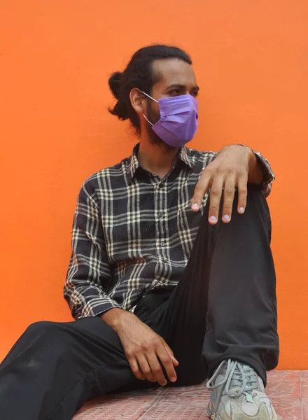 Front view of a young guy with hair bun wearing protective face mask while sitting outside, posing against orange wall with looking sideways — Stok fotoğraf