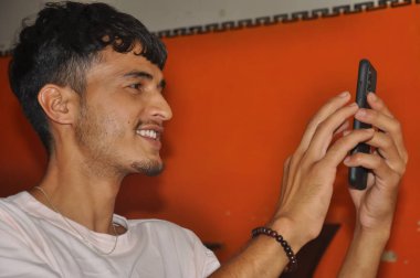 Kapalı mekanda otururken telefonunu kullanırken gülümseyen genç bir adamın yakın görüntüsü.