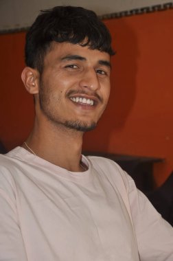 İyi görünümlü Hintli genç bir adamın yakın plan fotoğrafı. Gülümsüyor, evde oturuyor ve kameraya bakıyor.