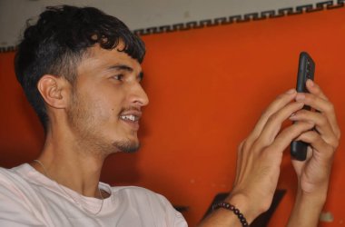 Kapalı mekanda otururken telefonunu kullanırken gülümseyen genç bir adamın yakın görüntüsü.