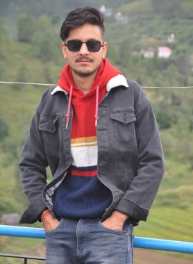 Yakışıklı, güneş gözlüğü takan, elleri cebinde poz veren ve dışarıda kameraya bakan Güney Asyalı bir genç. 