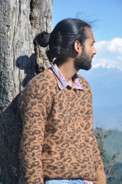 Saçları topuz gibi olan yakışıklı genç bir adam ağaçların karşısında dikilirken gözlerini kaçırıyor. 