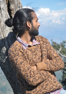 Saçları topuzlu genç bir adam kollarını kavuşturup ağaca yaslanırken yana bakıyor.  