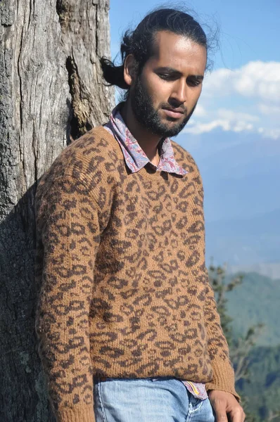 Doğada ağaca yaslanmış Güney Asyalı genç bir adam aşağıya bakıyor. 