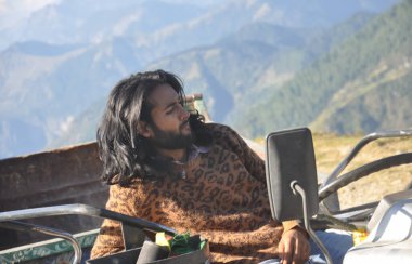 Güzel görünümlü, uzun saçlı genç bir erkek manken, traktörün üzerinde otururken yana bakıyor.