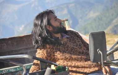 Güzel görünümlü, uzun saçlı genç bir erkek manken, traktörün üzerinde otururken yana bakıyor.