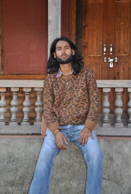 Eski tapınakta otururken kameraya bakan uzun saçlı seksi bir genç adam.